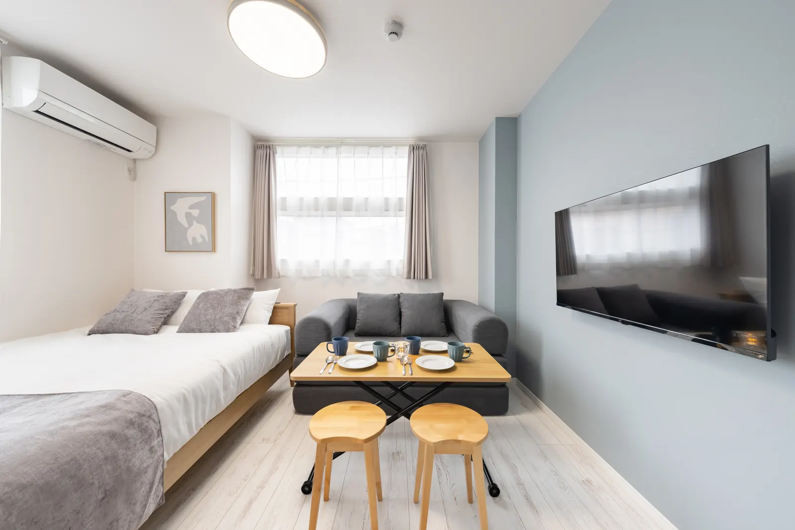 Studio unit at Intervia Horikiri II with a double bed, wooden dining table with stools, grey sofa bed and wall-mounted TV against a pale blue wall